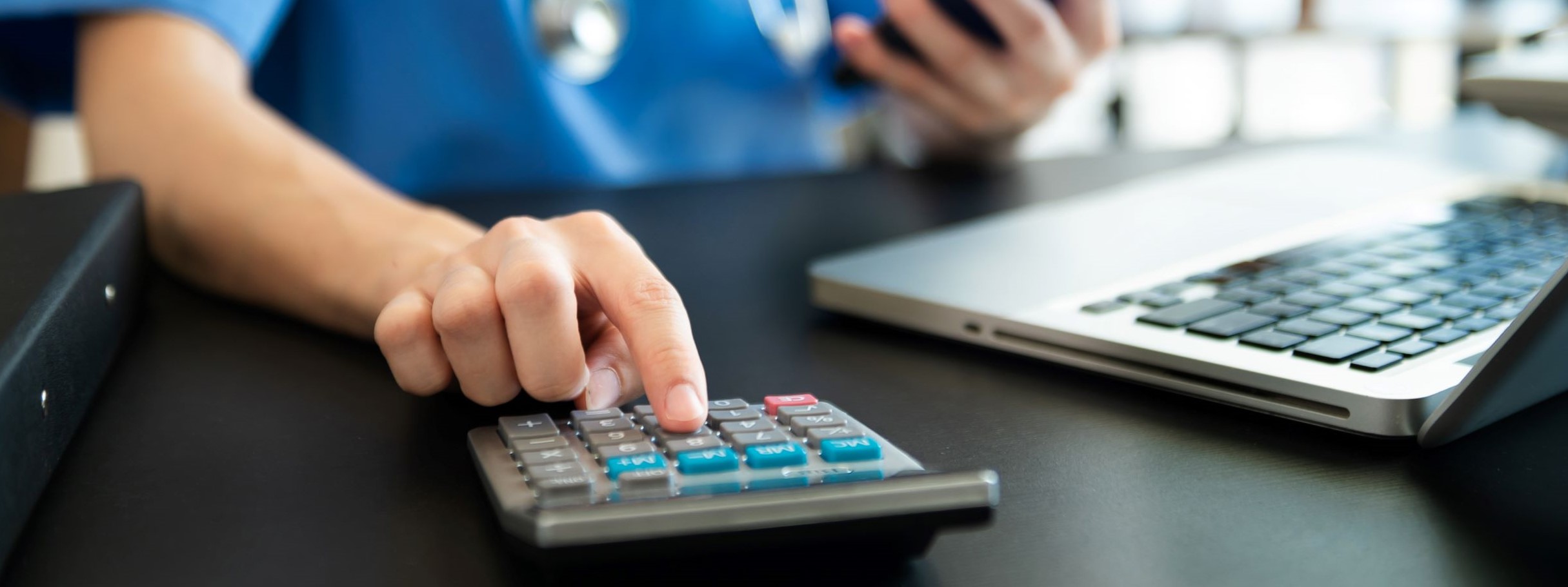 An individual in scrubs with a stethoscope operates a calculator with one hand while holding a phone in the other  