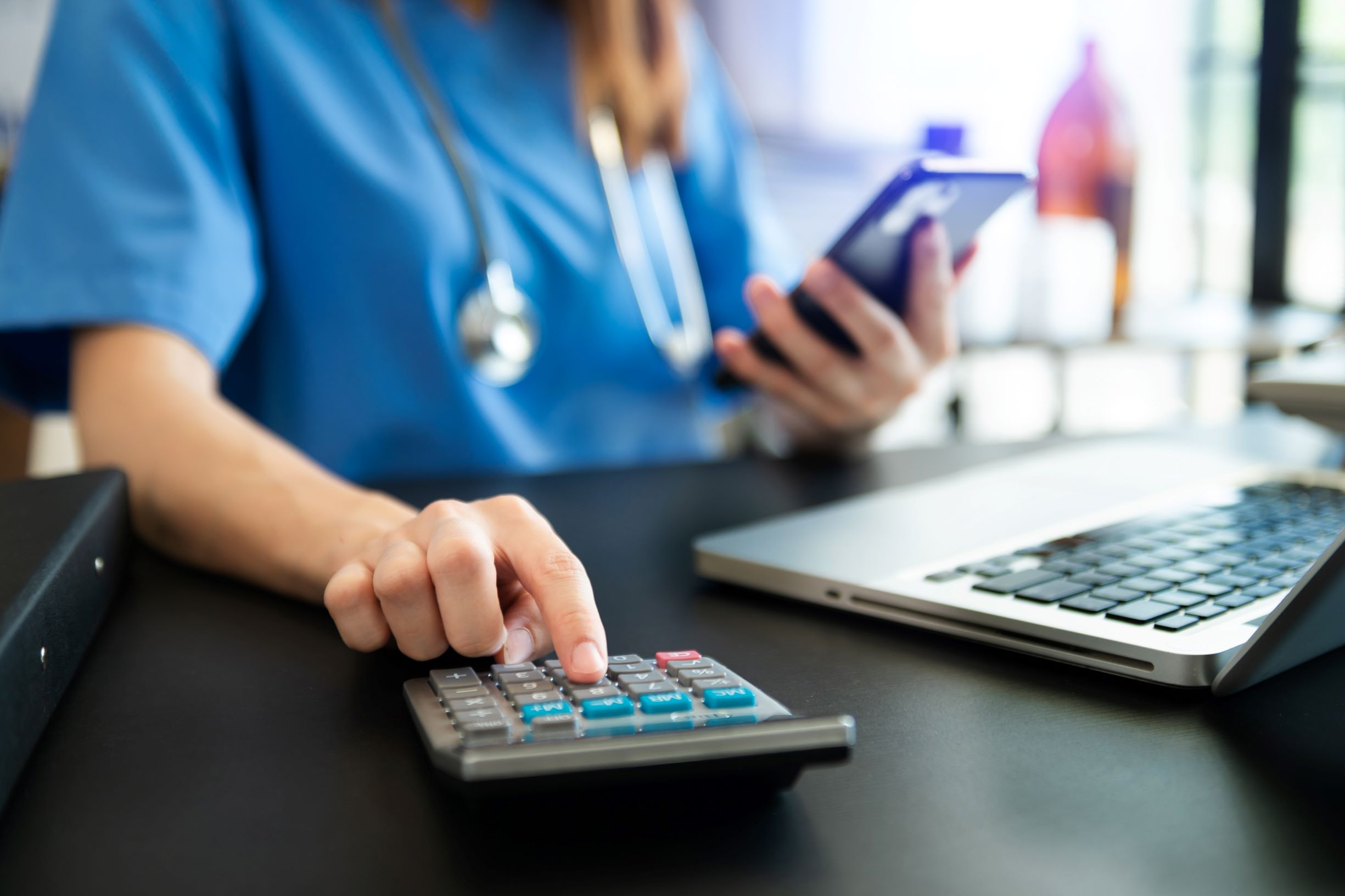 An individual in scrubs with a stethoscope operates a calculator with one hand while holding a phone in the other  