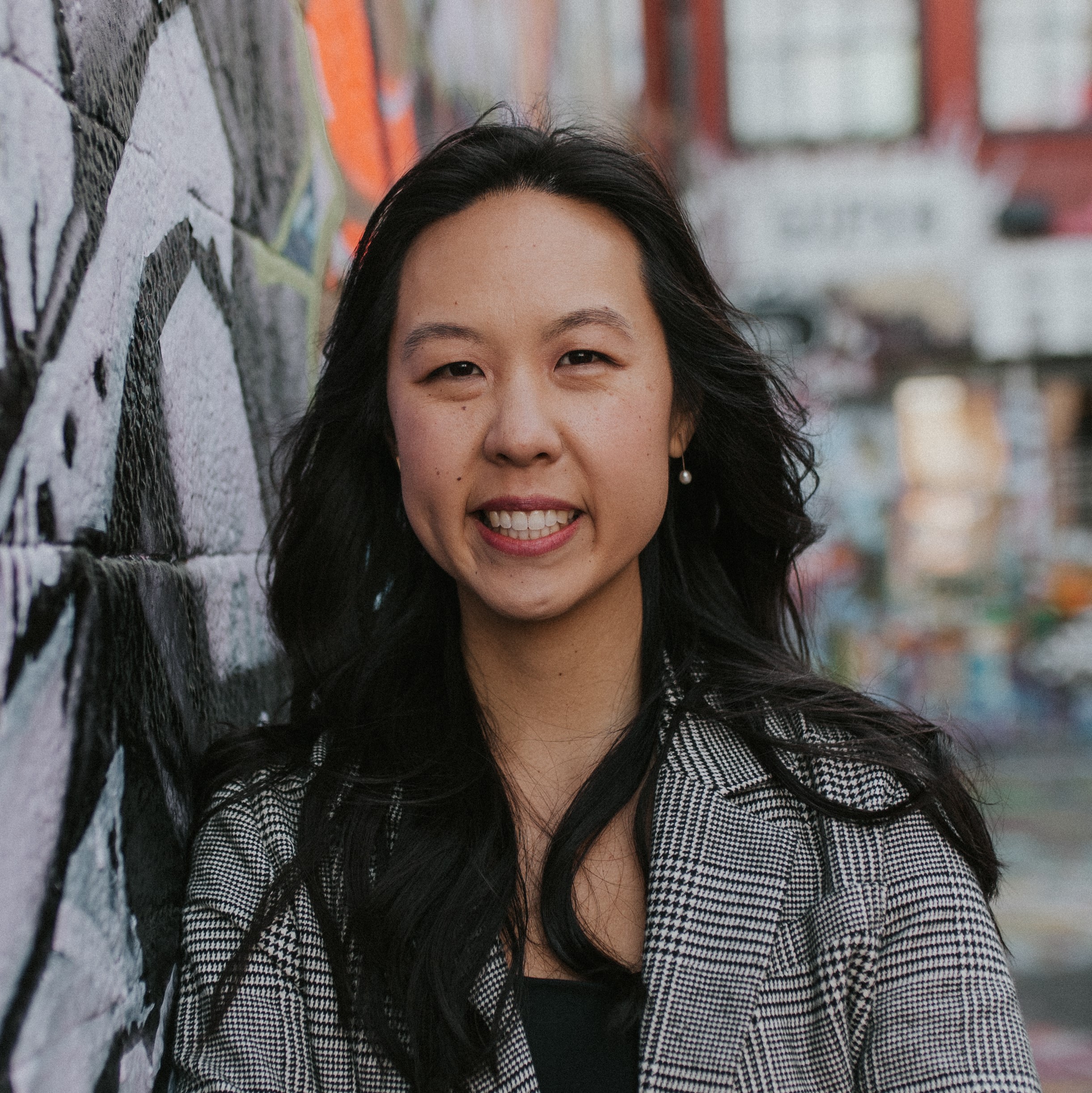 Cochlear Center faculty member Alison Huang headshot. Huang is wearing a black and white ensemble and is leaning on a graffiti-covered wall 