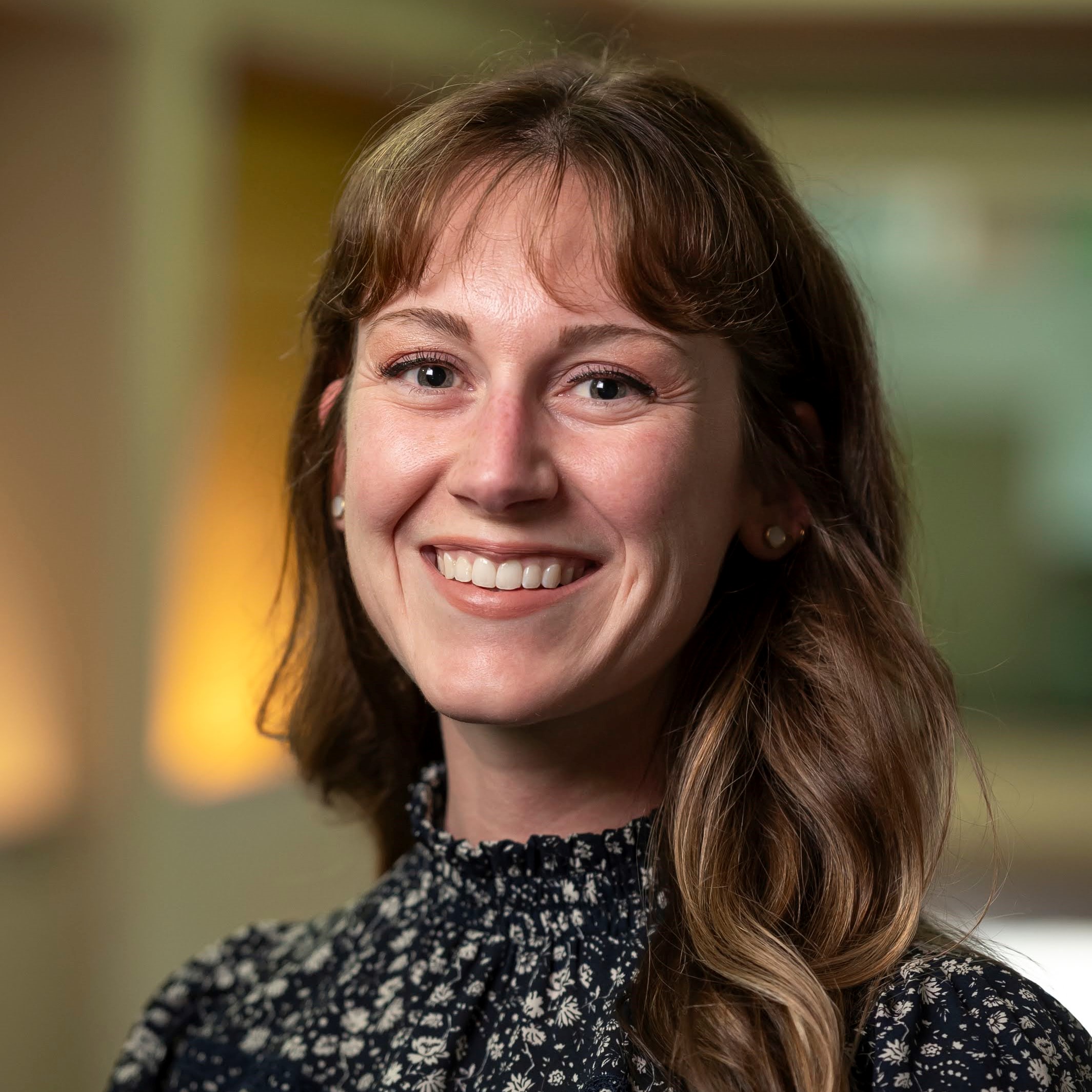 Faculty audiologist Clarice Myers smiles at the viewer. She has long hair with bangs, and is wearing a floral top with a high neck