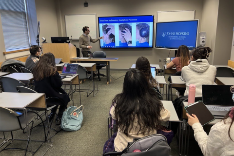 Photo of various students in a classroom in the foreground, listening to the profesor giving the class in the background